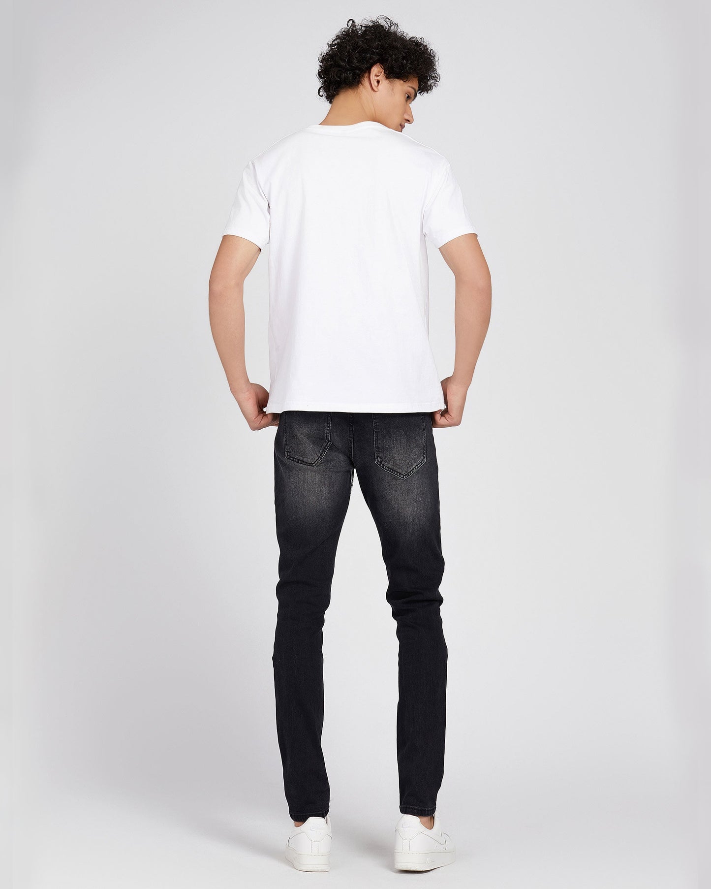 Man wearing Logeqi black ripped graffiti jeans with white t-shirt and sneakers viewed from back