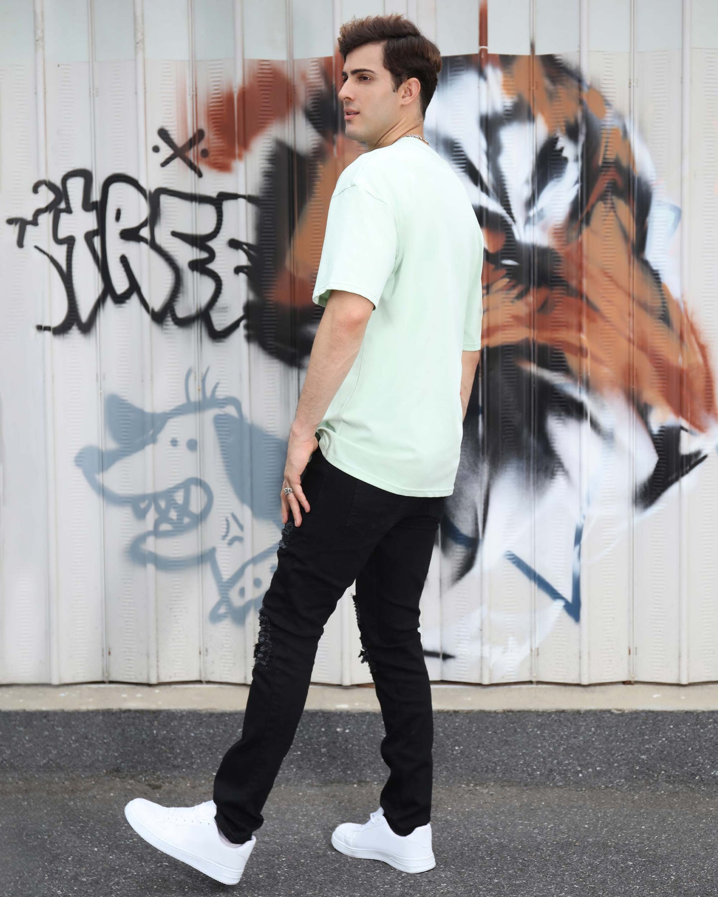 Man wearing Logeqi black ripped jeans for men with a mint green t-shirt against a graffiti background