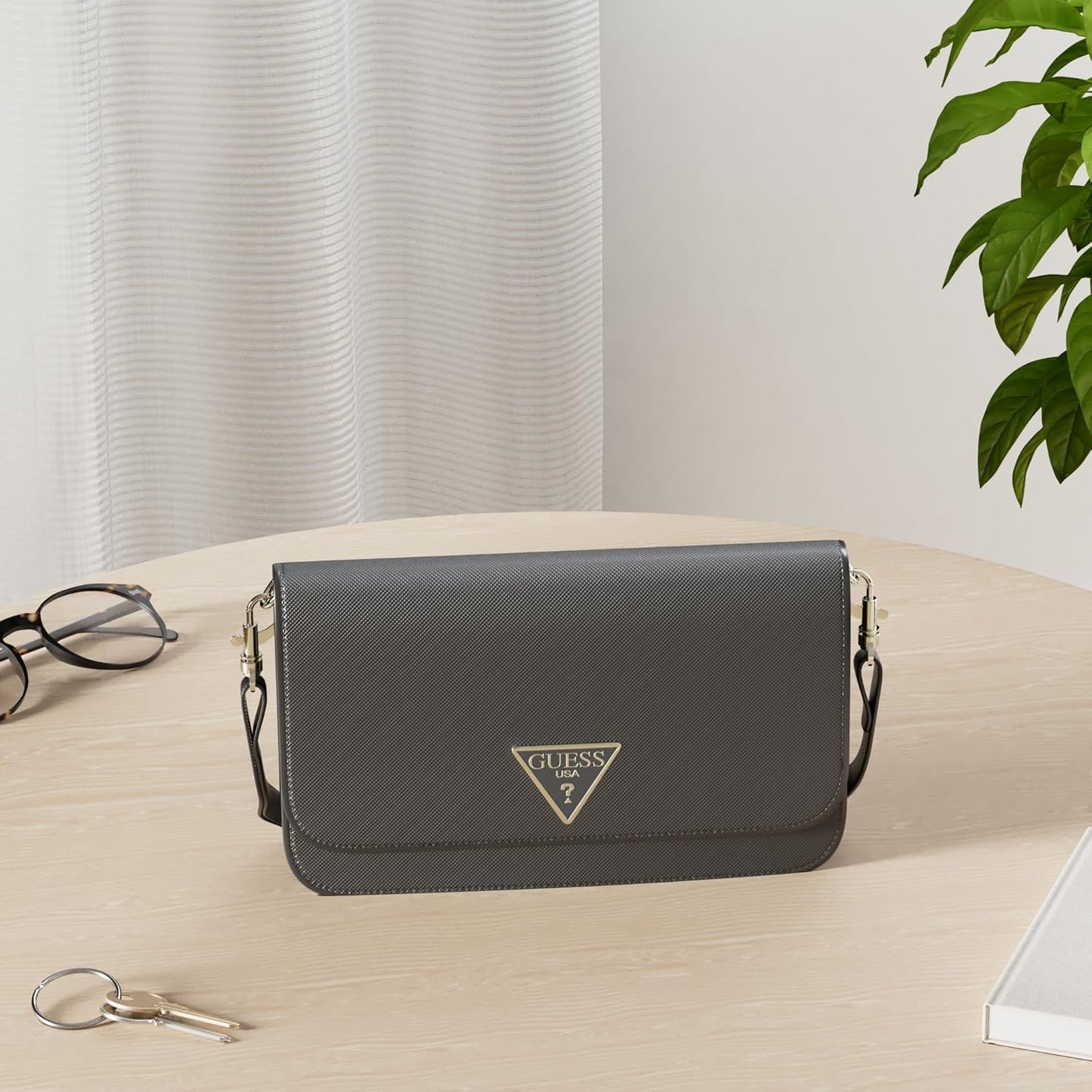 GUESS Noelle crossbody bag in gray placed on a table with stylish accessories