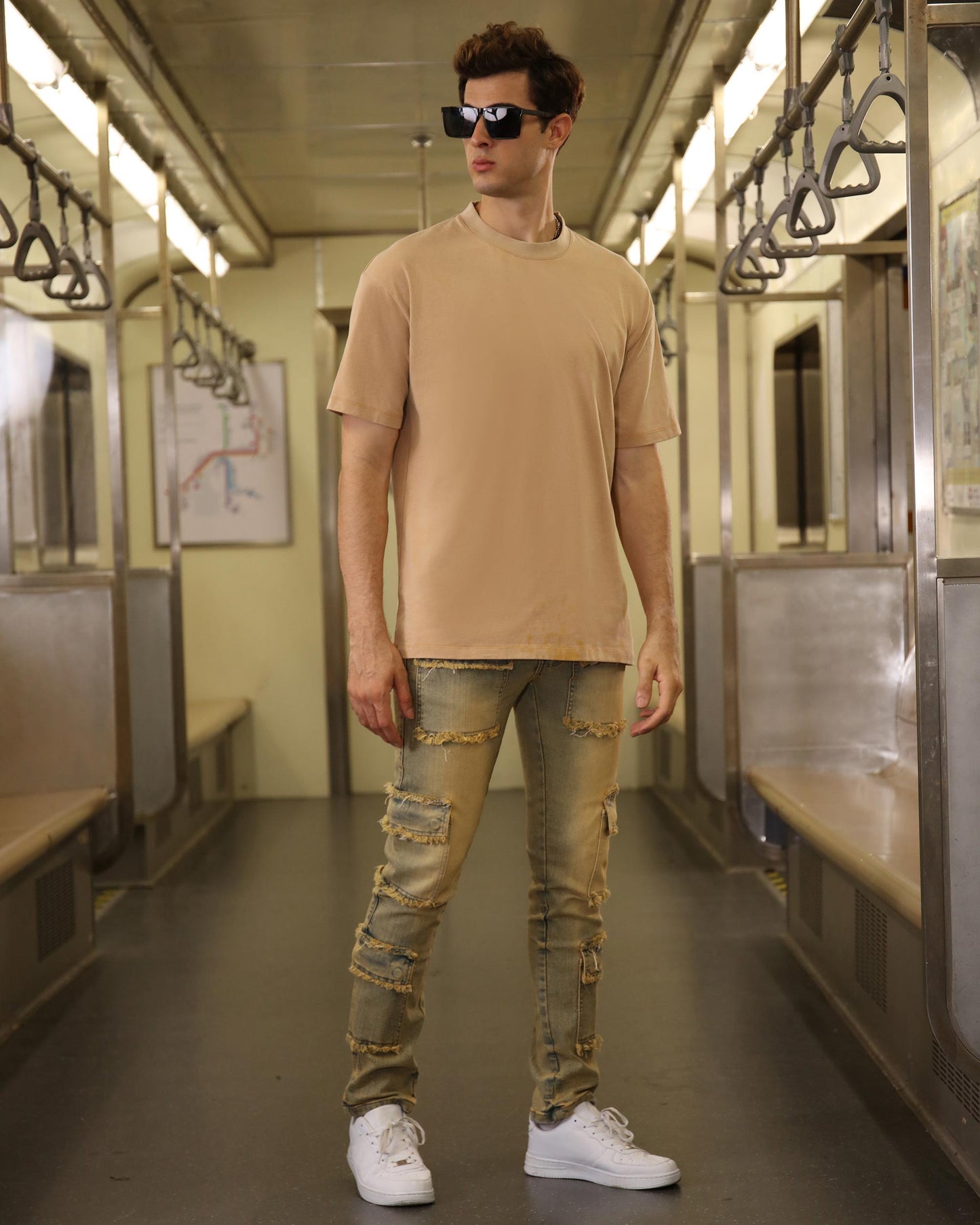 Man wearing Logeqi raw edge brown jeans with distressed details and white sneakers standing in subway car