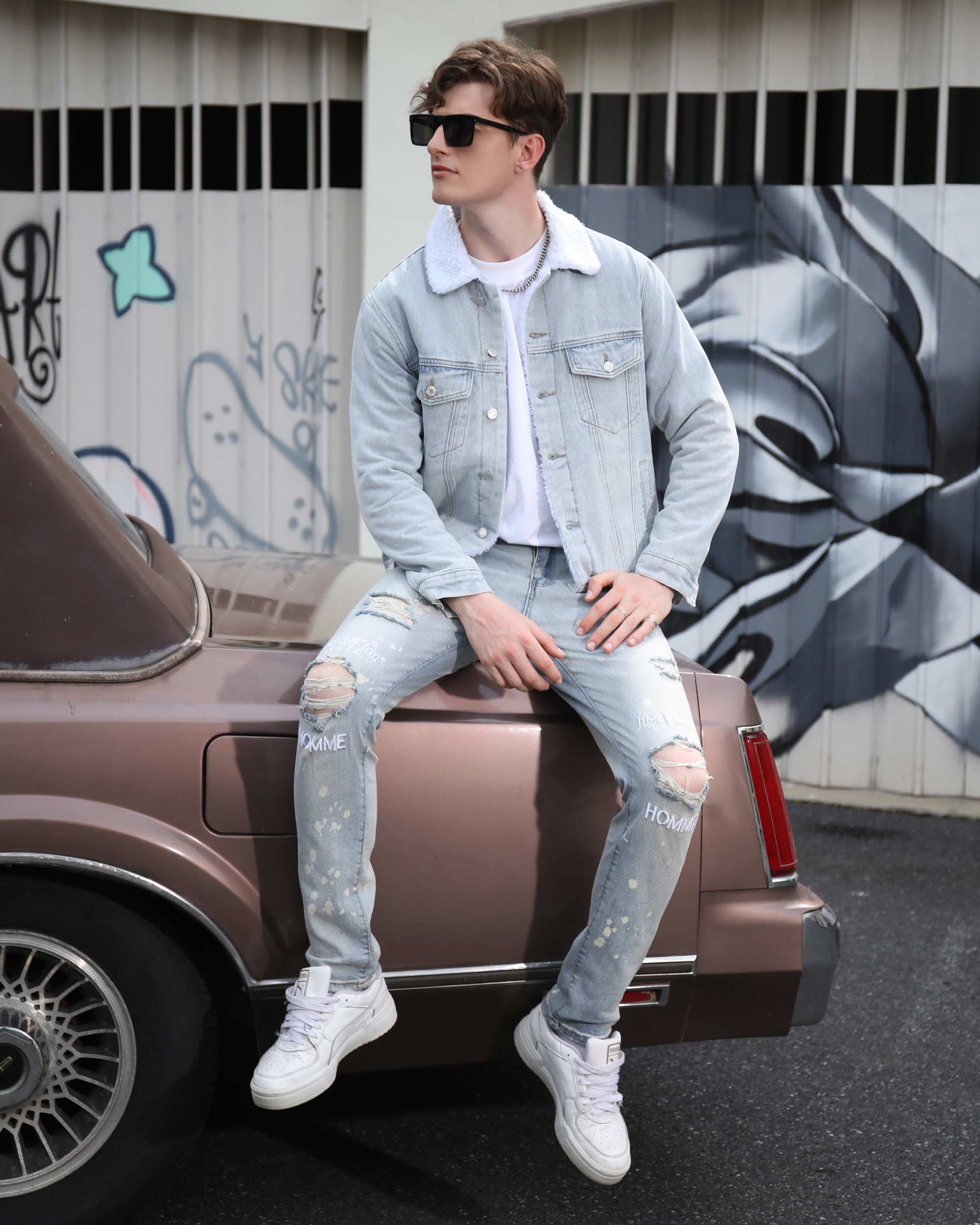 Man wearing spray painted light blue ripped jeans by Logeqi sitting on vintage car in urban setting