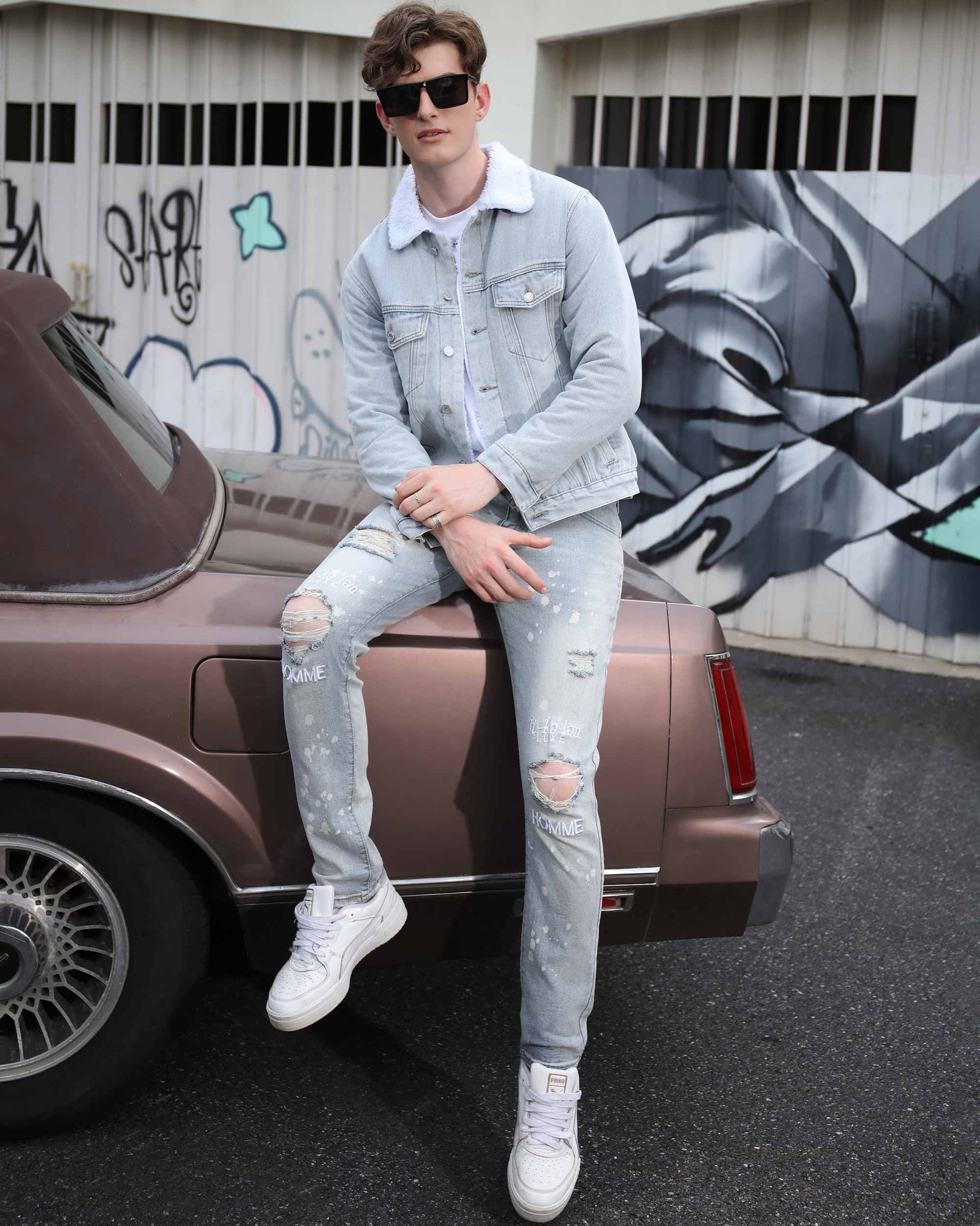 Man wearing spray painted light blue ripped jeans sitting on car bumper with denim jacket and white sneakers