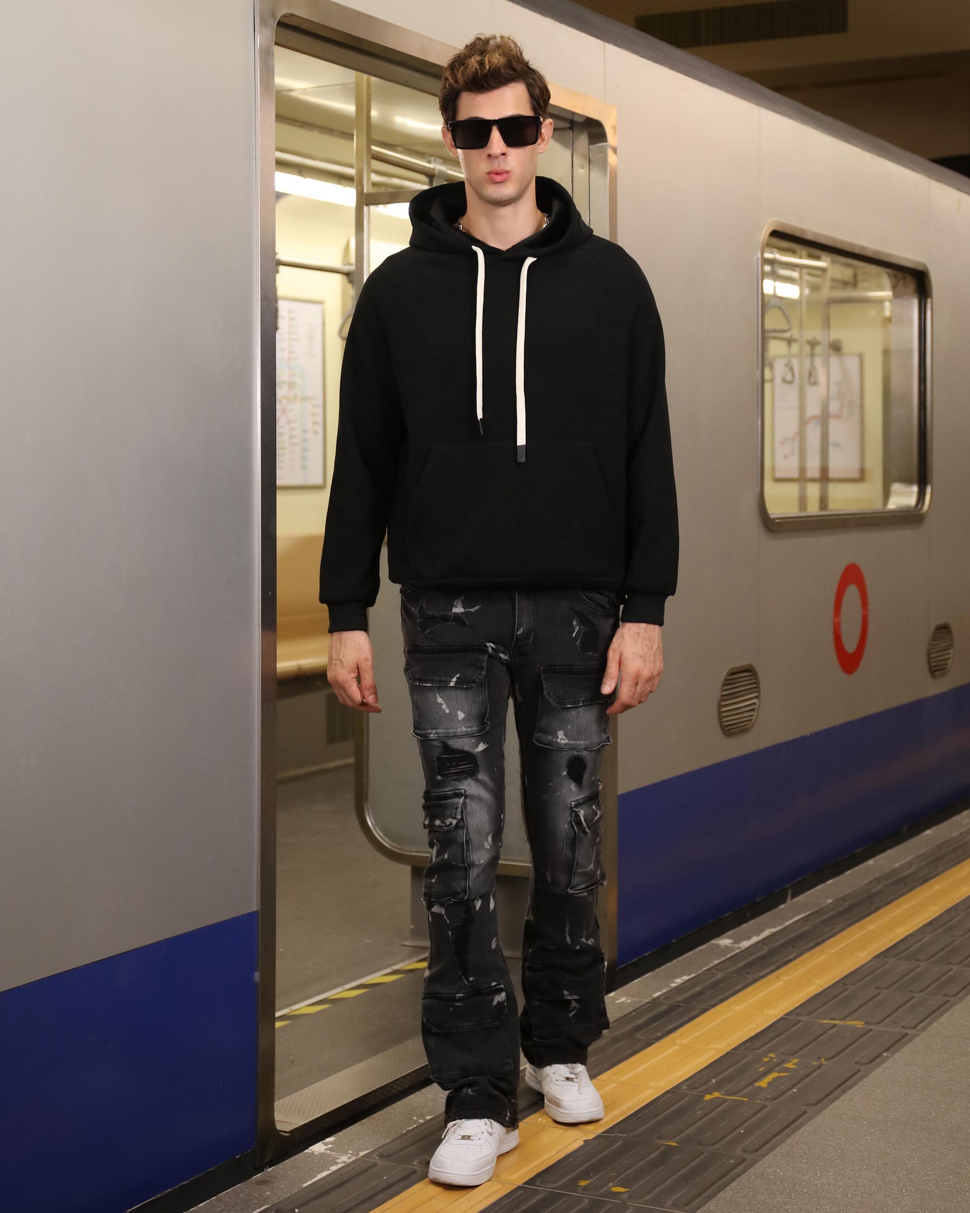 Man wearing distressed black cargo jeans by Logeqi paired with black hoodie and white sneakers at subway station
