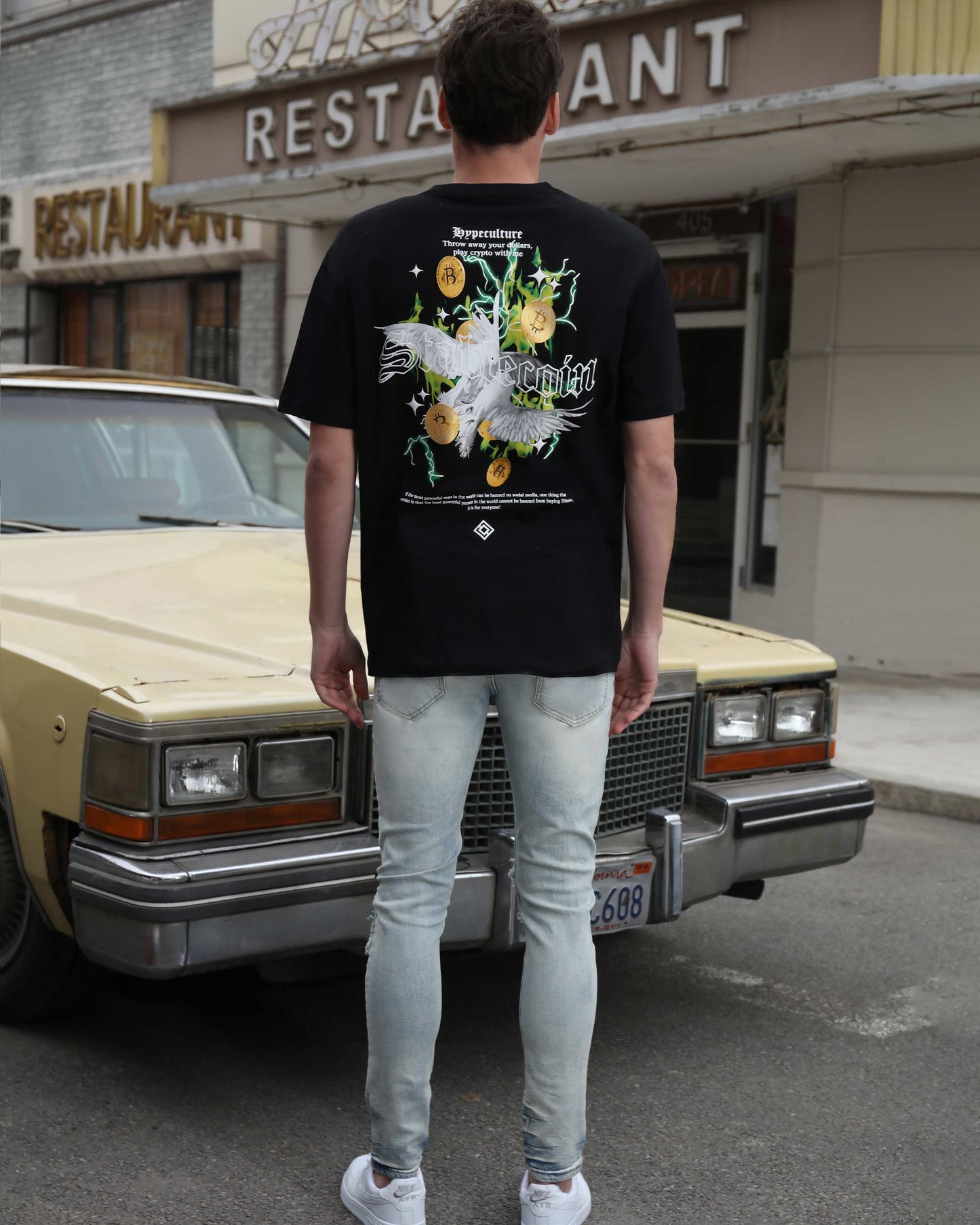 Man wearing Logeqi light wash graffiti jeans paired with a black graphic t-shirt standing by a vintage car
