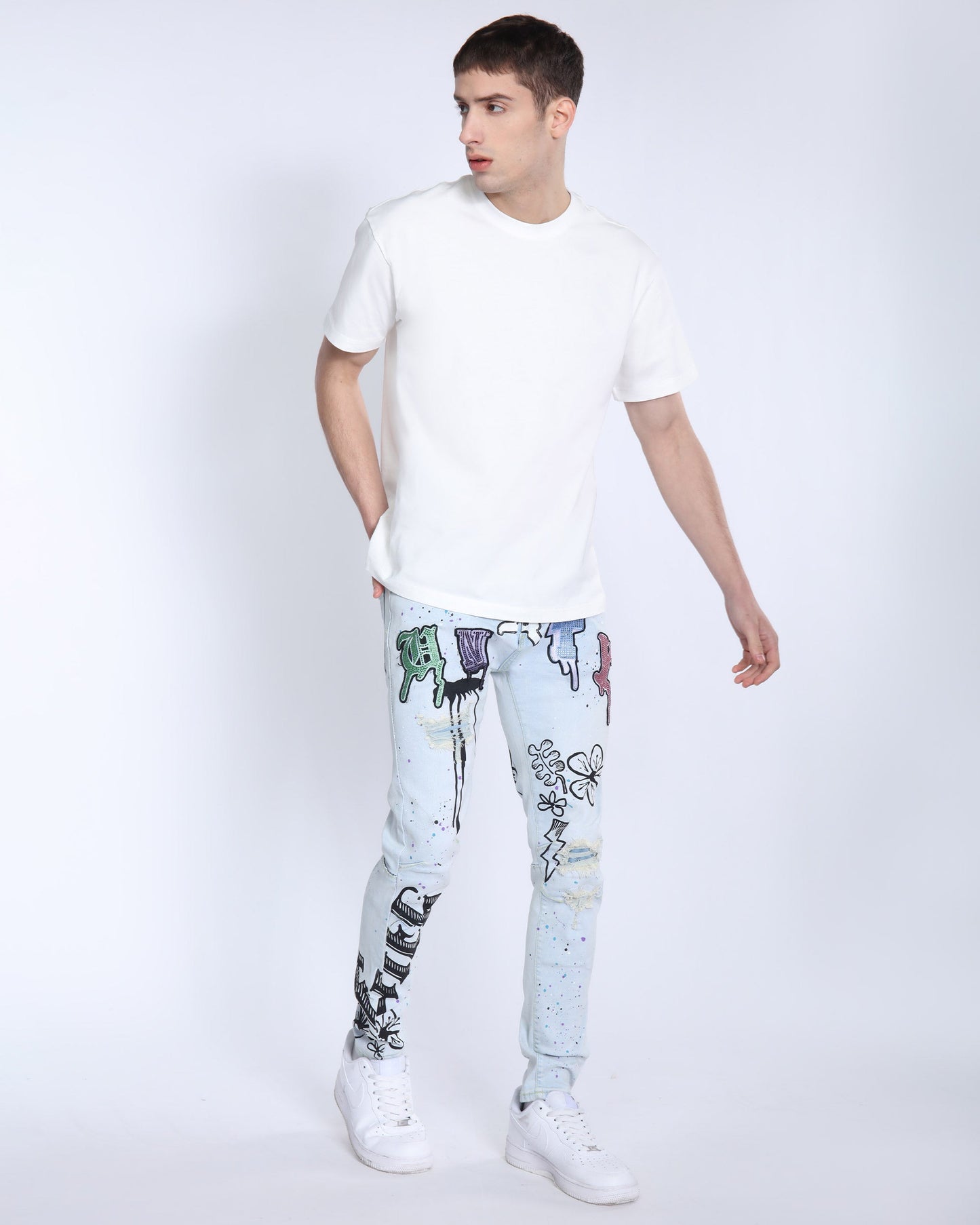 Man wearing Logeqi comic painted ripped jeans with colorful graffiti designs paired with a white t-shirt and white sneakers